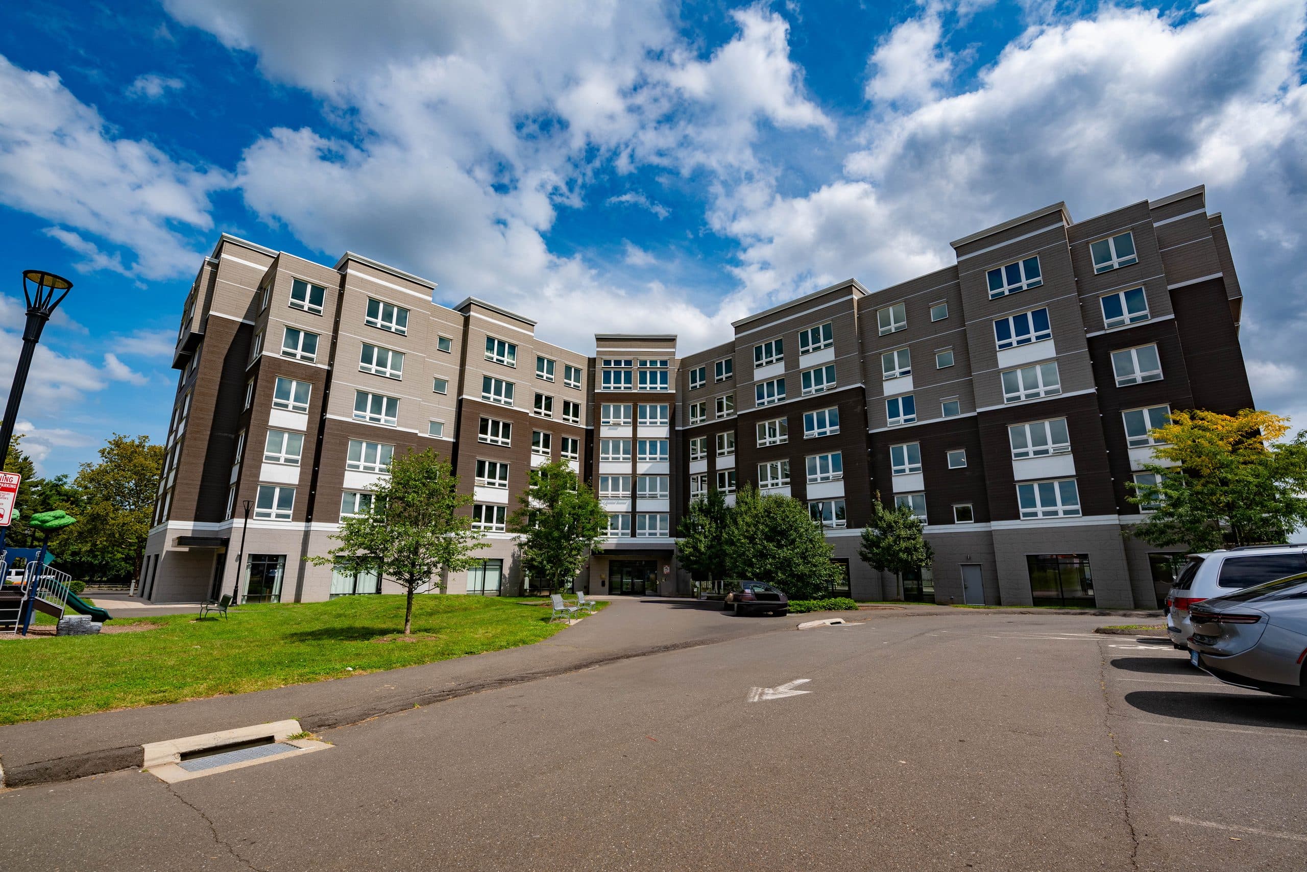Columbus Commons — a six-storey apartment building in charcoal and grey masonry, seen across its front lawn under a bright summer sky