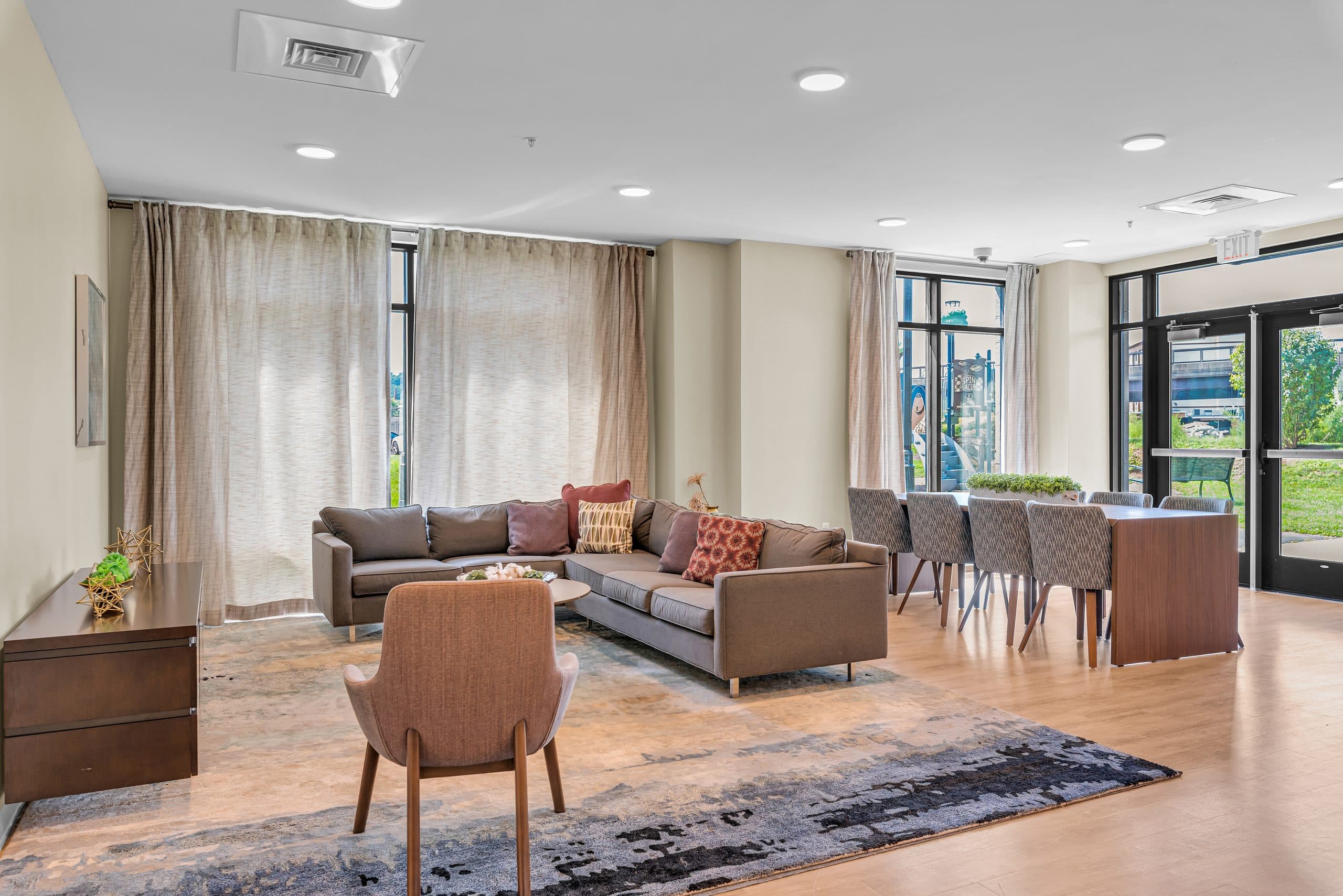 The Columbus Commons resident lounge — a sectional and armchair on a patterned rug, a long dining table beyond, and floor-to-ceiling windows along two walls