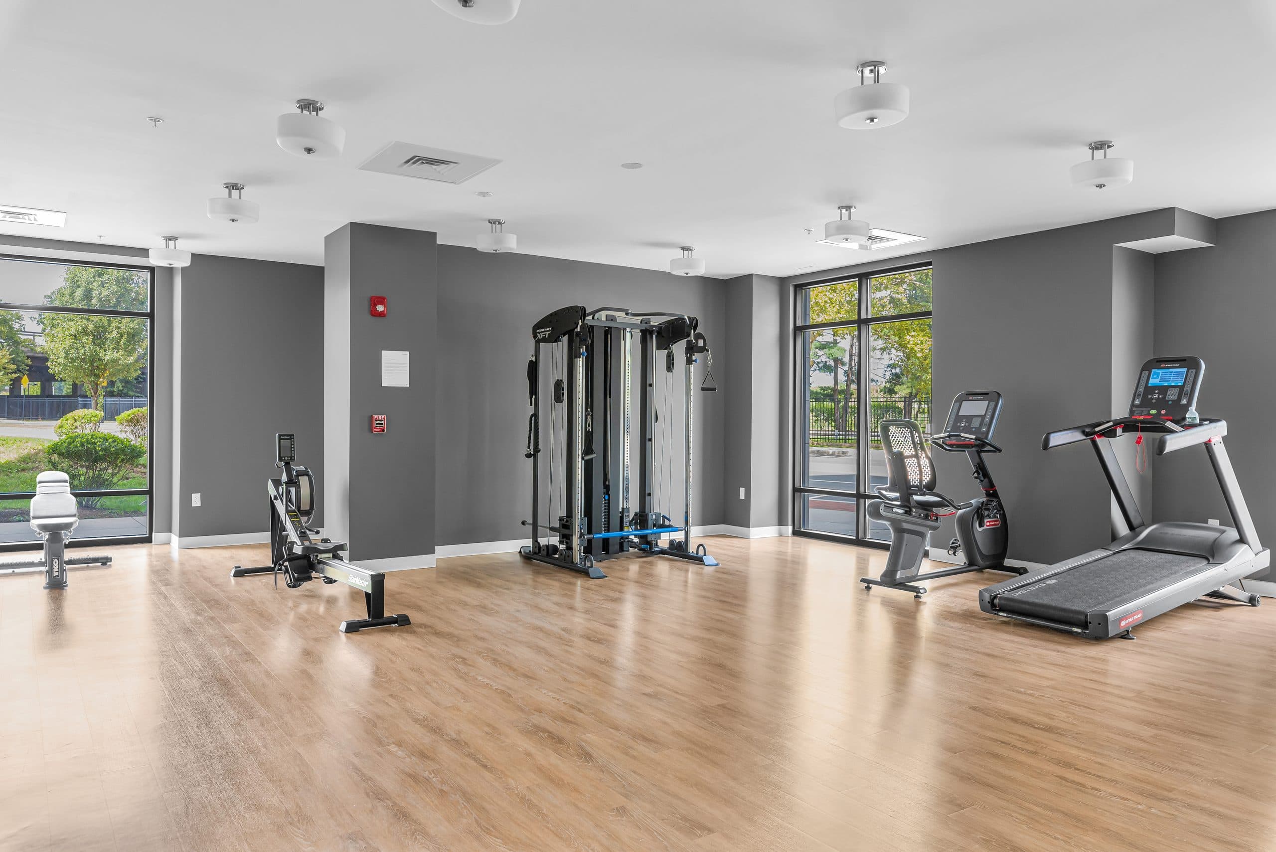 The Columbus Commons fitness center — a treadmill, recumbent bike, rower, and functional trainer on pale wood flooring beside floor-to-ceiling windows